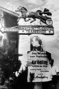 Beschriftung des Wegweisers: „Fußweg Gaststätte und Gestüt Traberhof“ – Text auf Plakat: „Das Wunder von Harlaching. Gröning heilt auf der Straße. Experiment missglückt – Heilungen gelungen. Lesen Sie Abendzeitung“